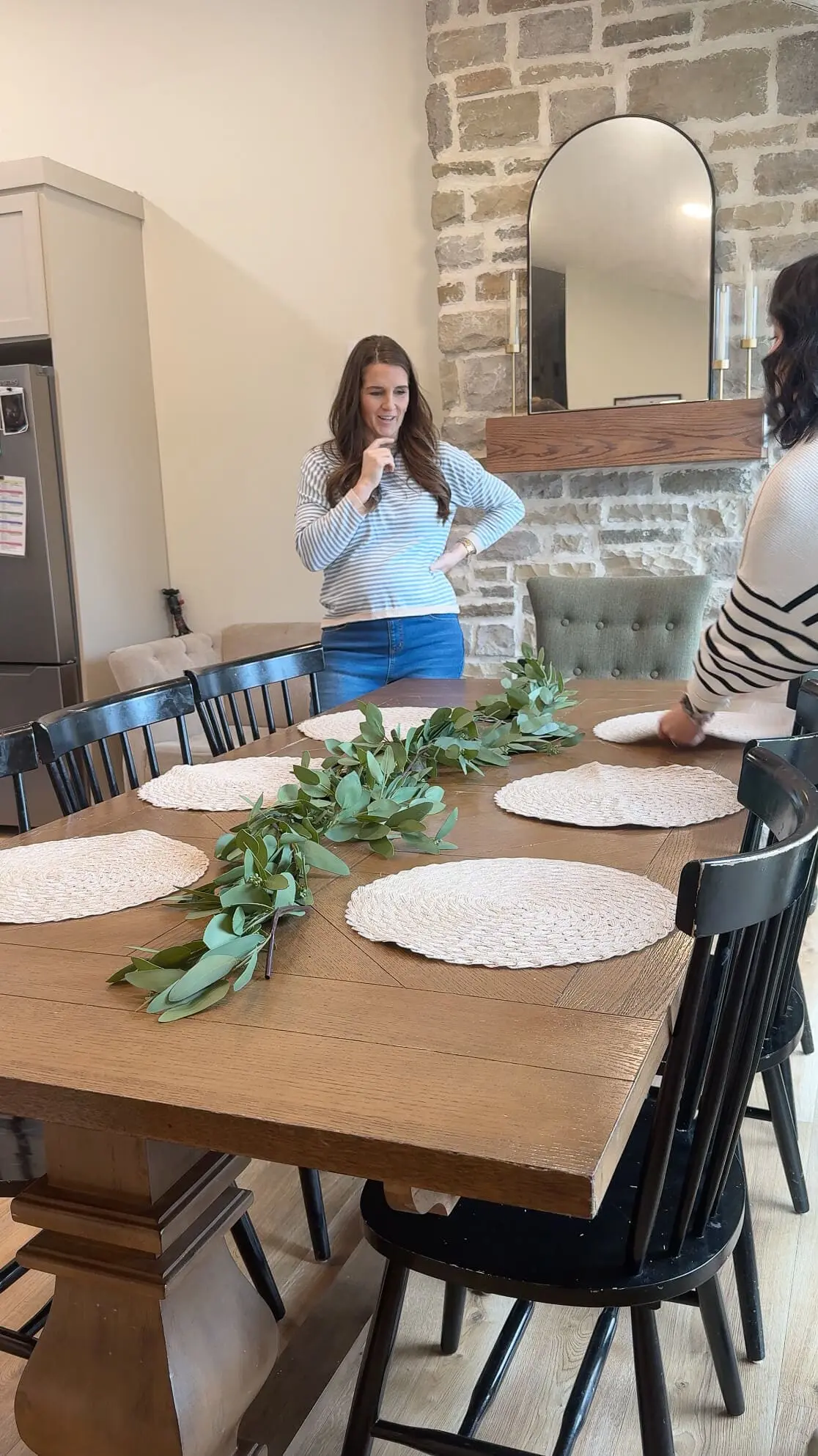 A dining table in the process of being styled, with a centerpiece of eucalyptus garland running down the middle. A woman in a striped top and jeans is thoughtfully planning the arrangement while woven placemats are being laid out.